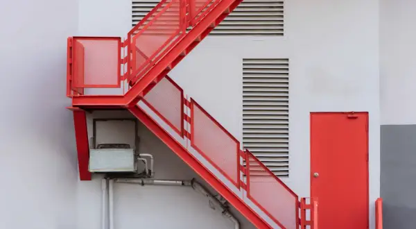 Czerwone metalowe schody zewnętrzne z siatkowymi barierkami obok białej ściany z kratkami wentylacyjnymi i drzwiami.